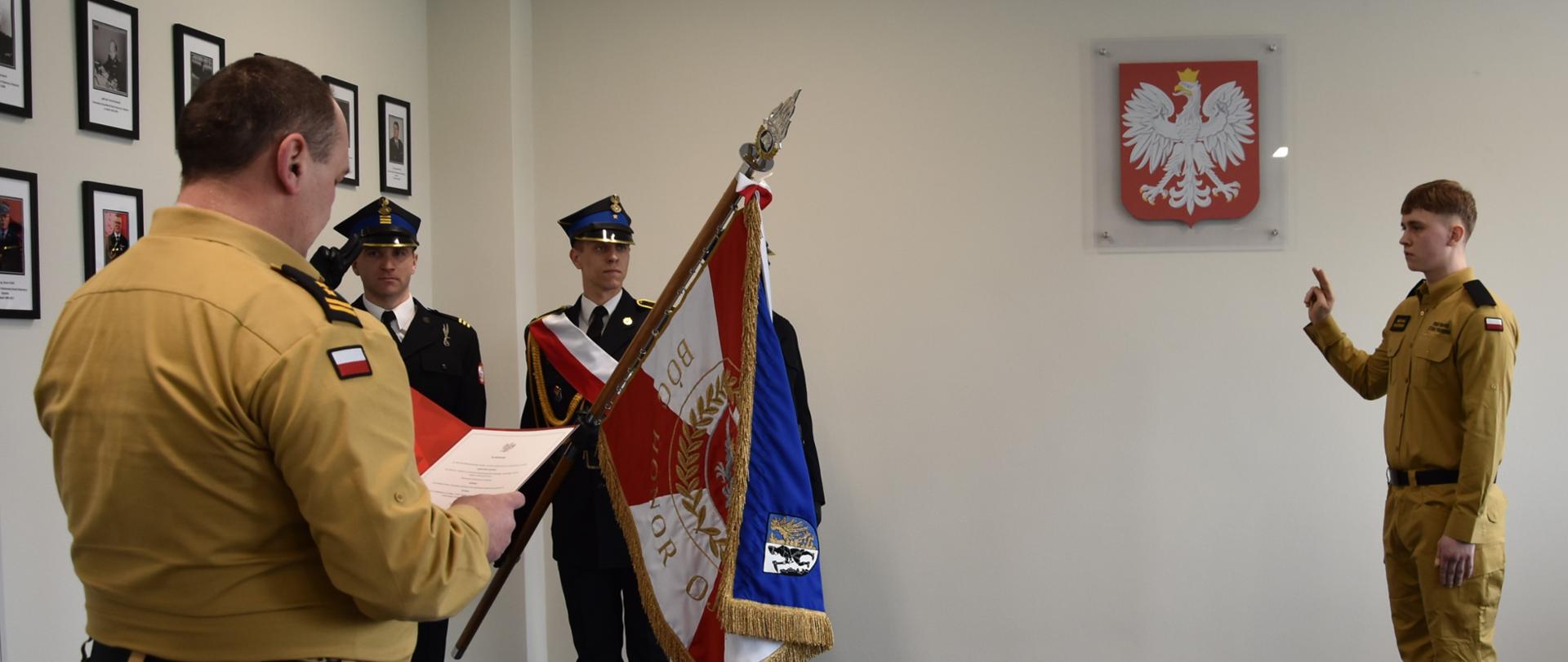 Moment ślubowania, na pierwszym planie strażak trzymający teczkę, dalej poczet sztandarowy i strażak składający ślubowanie. Na ścianie godło.