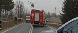 Kolorowe zdjęcie wykonane w pochmurny dzień przedstawia drogę, na której stoją częściowo widoczne czerwone pojazdy straży pożarnej i karetka pogotowia ratunkowego. Nad pojazdami widać żółty śmigłowiec Lotniczego Pogotowia Ratunkowego. Z boku widać uszkodzony pojazd osobowy i umundurowanych strażaków. Po bokach widoczne szare drzewa.