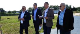 Wiceminister sportu i turystyki Piotr Borys oraz trzej inni mężczyźni idą asfaltową alejką. Wzdłuż alejki rośnie trawa. W tle widać zabudowania.