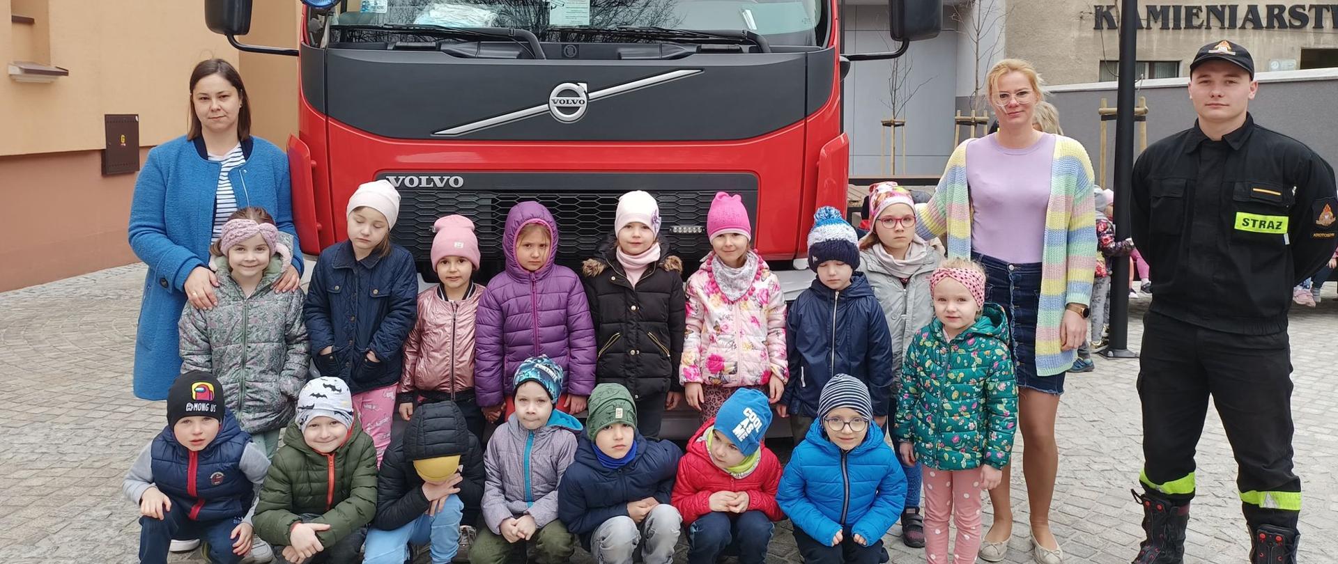 Zdjęcie grupowe uczestników pogadanki ze strażakami. Na tle pojazdu pożarniczego na placu szkolnym stoi grupa dzieci z opiekunkami oraz strażak uczestniczący w szkoleniu