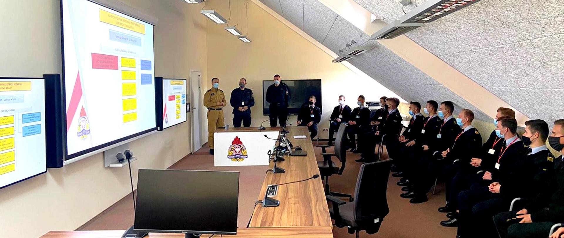 Podchorążowie siedzą w pomieszczeniu podczas wykładu, funkcjonariusz prowadzący zajęcia stoi na wprost obok niego stoi dwóch strażaków , po lewo ściana z monitorami