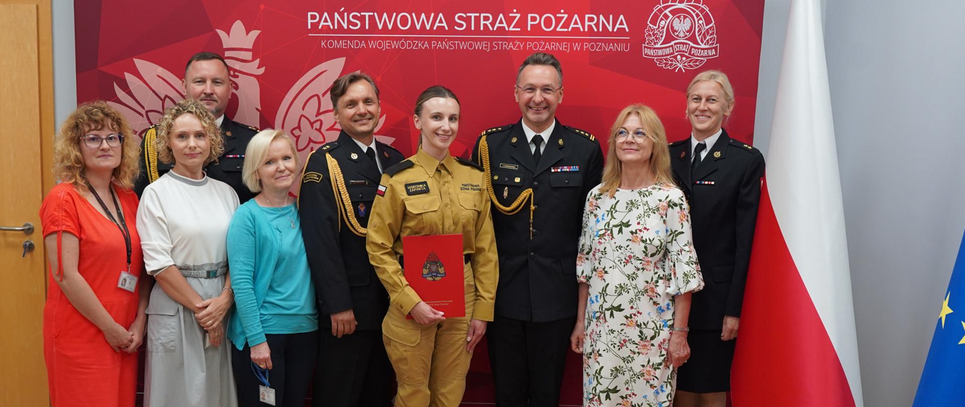 Grupa uczestników uroczystości pozuje do wspólnego zdjęcia na tle ścianki Komendy Wojewódzkiej Państwowej Straży Pożarnej w Poznaniu. W centrum stoi strażaczka w mundurze służbowym, trzymająca czerwoną teczkę z dokumentami, otoczona przez funkcjonariuszy w mundurach galowych oraz osoby cywilne. Po prawej stronie widoczne są flagi Polski i Unii Europejskiej.