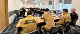 Na zdjęciu widzimy salę wykładową, a w niej strażaków: słuchaczy w tle w czarnych uniformach oraz wykładowców na pierwszym planie tyłem do zdjęcia z napisem na piaskowych mundurach Państwowa Straż Pożarna