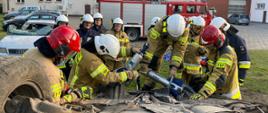Ubrani w ubrania specjalne strażacy stoją przy przewróconym na dach aucie, jeden z nich rozcina podwozie za pomocą nożyc hydraulicznych 