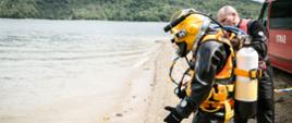 Na zdjęciu strażak nurek ubrany w skafander i hełm przeznaczony do pracy w wodach skażonych i zanieczyszczonych stojący na brzegu jeziora Solińskiego, za nim nurek instruktor pomagający podczas wejścia do wody