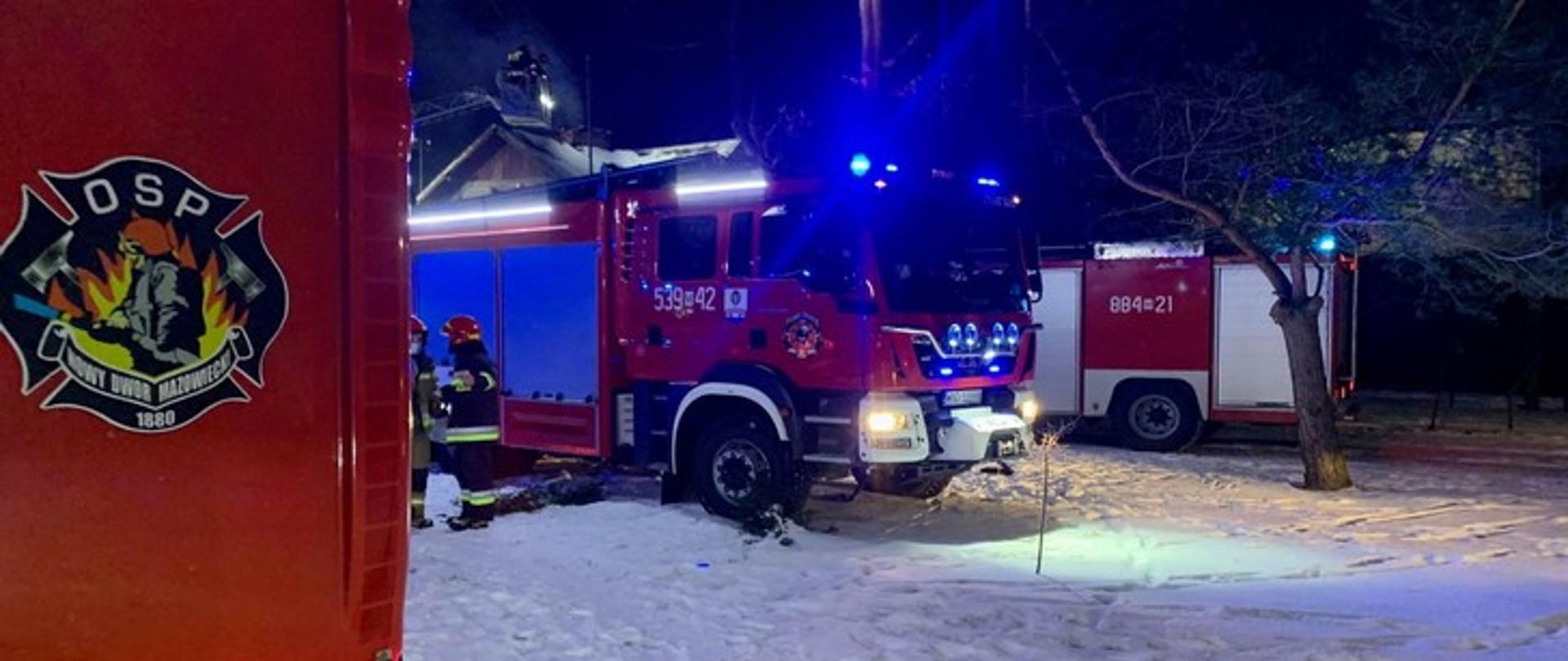 Zdjęcie w warunkach nocnych przedstawia dwa samochody pożarnicze z włączonymi niebieskimi światłami ostrzegawczymi, z lewej strony dwóch strażaków.