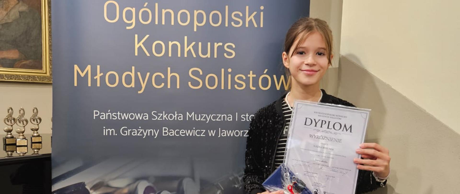 Dziewczynka stoi przed banerem Konkursu Młodych Solistów w Jaworznie i trzyma w rękach dyplom oraz nagrodę.