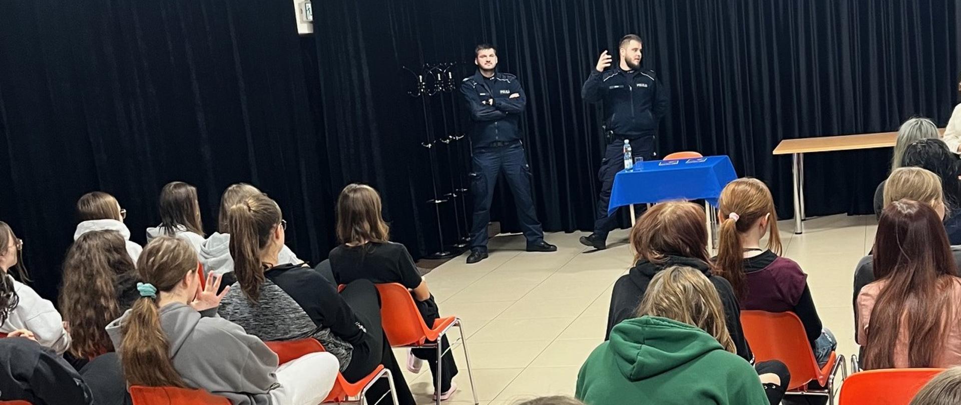 W oddali na tle czarnej kurtyny stoją dwaj umundurowani policjanci, jeden z nich patrzy w lewą stronę i unosi prawą dłoń. Przed nimi stolik z niebieskim obrusem i butelką wody. W pierwszym planie widać od tyłu kilkunastu młodych ludzi siedzących na pomarańczowych krzesłach.