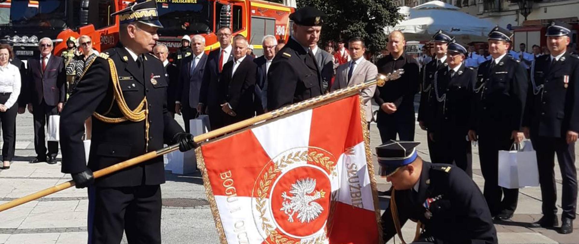 Zastępca KG PSP nachyla sztandar, Komendant Miejski klęcząc całuje sztandar, w tle uczestnicy