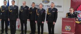 Od lewej strony stoi sześciu oficerów PSP, po prawej stronie oficer PSP przemawiający z mównicy