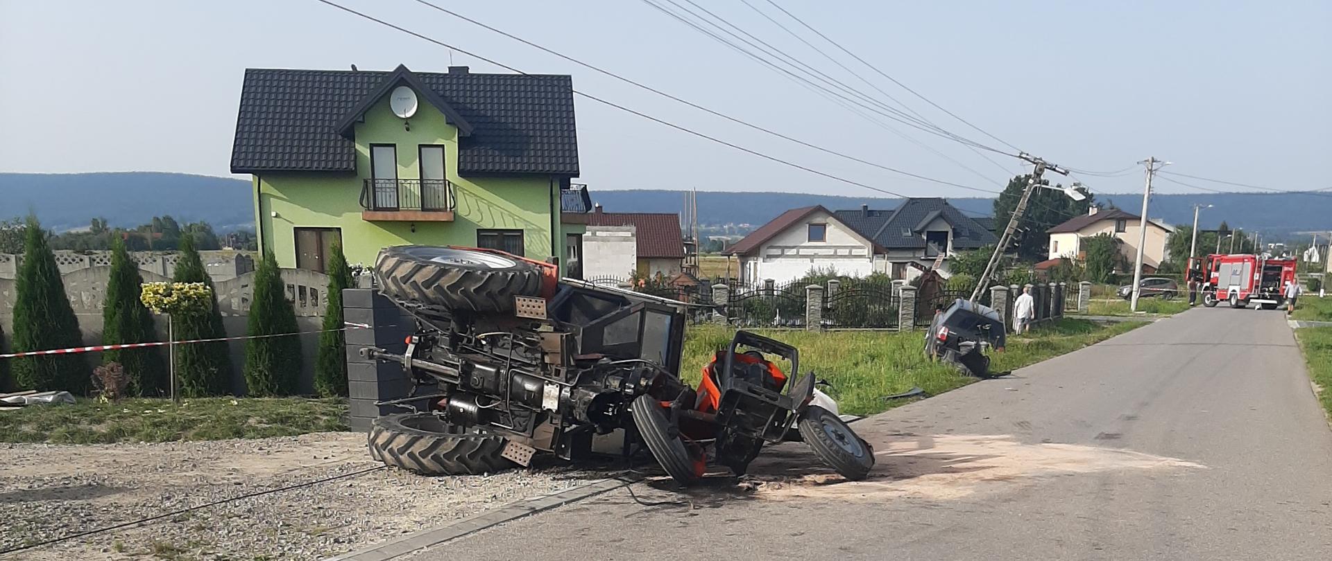 Zdjęcie przedstawia drogę gminną na której doszło do zderzenia samochodu osobowego i ciągnika rolniczego. Po lewej stronie drogi widać kolejno: uszkodzony ciągnik, dalej auto osobowe i w tle samochody pożarnicze.