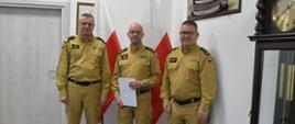Trzech funkcjonariuszy Państwowej Straży Pożarnej w mundurach koloru musztardowego stoi obok siebie strażak w środku trzyma białą teczkę za mężczyznami stoją dwie flagi Polski na ścianą wiszą obraz oraz szabla obok ustawiony jest zegar.