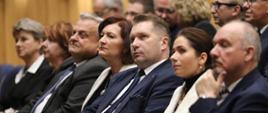Minister w otoczeniu grupy ludzi siedzi na sali, wszyscy czegoś z uwagą słuchają.