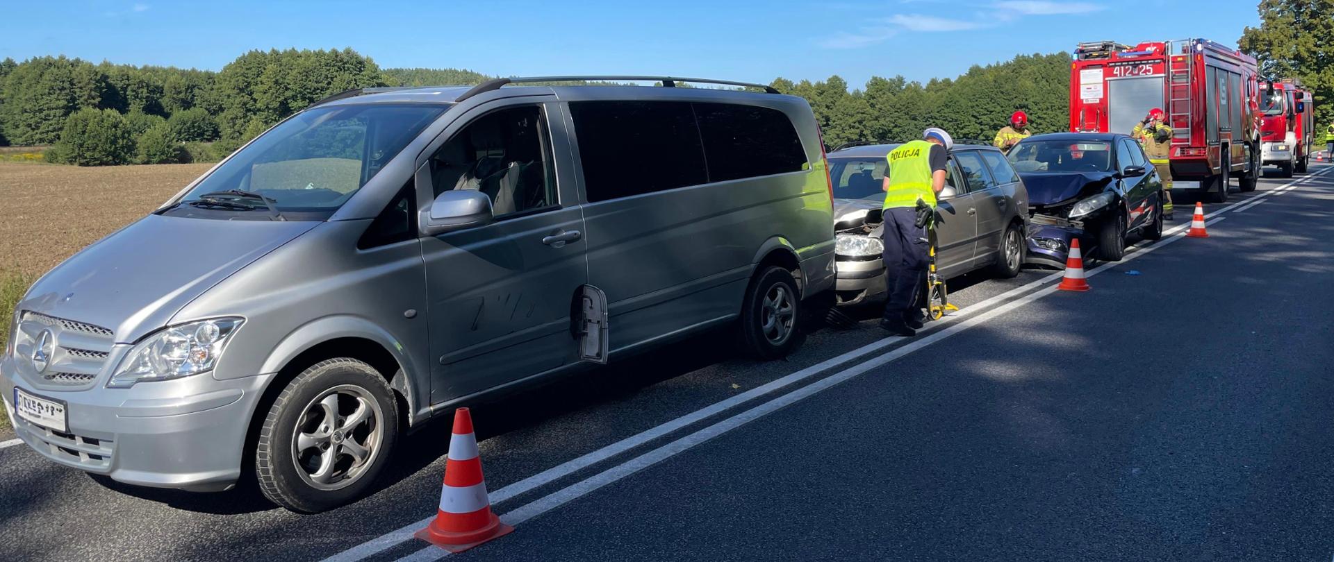 Kolumna złożona z trzech uszkodzonych pojazdów na jednej połowie jezdni na tle pojazdów Państwowej Straży Pożarnej.