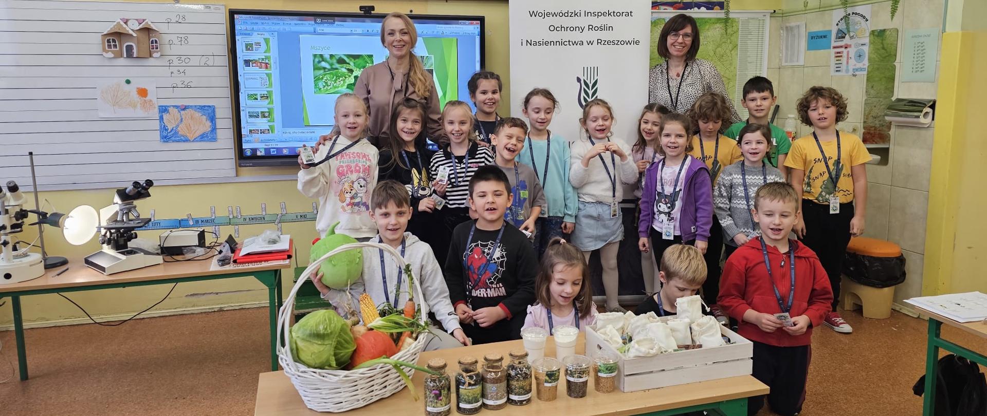 Grupa dzieci i dwoje dorosłych w szkolnej klasie, stoją w rogu sali przy ścianie. Wszyscy uśmiechają się do zdjęcia, chłopczyk trzyma w ręku zieloną okrągłą maskotkę. 