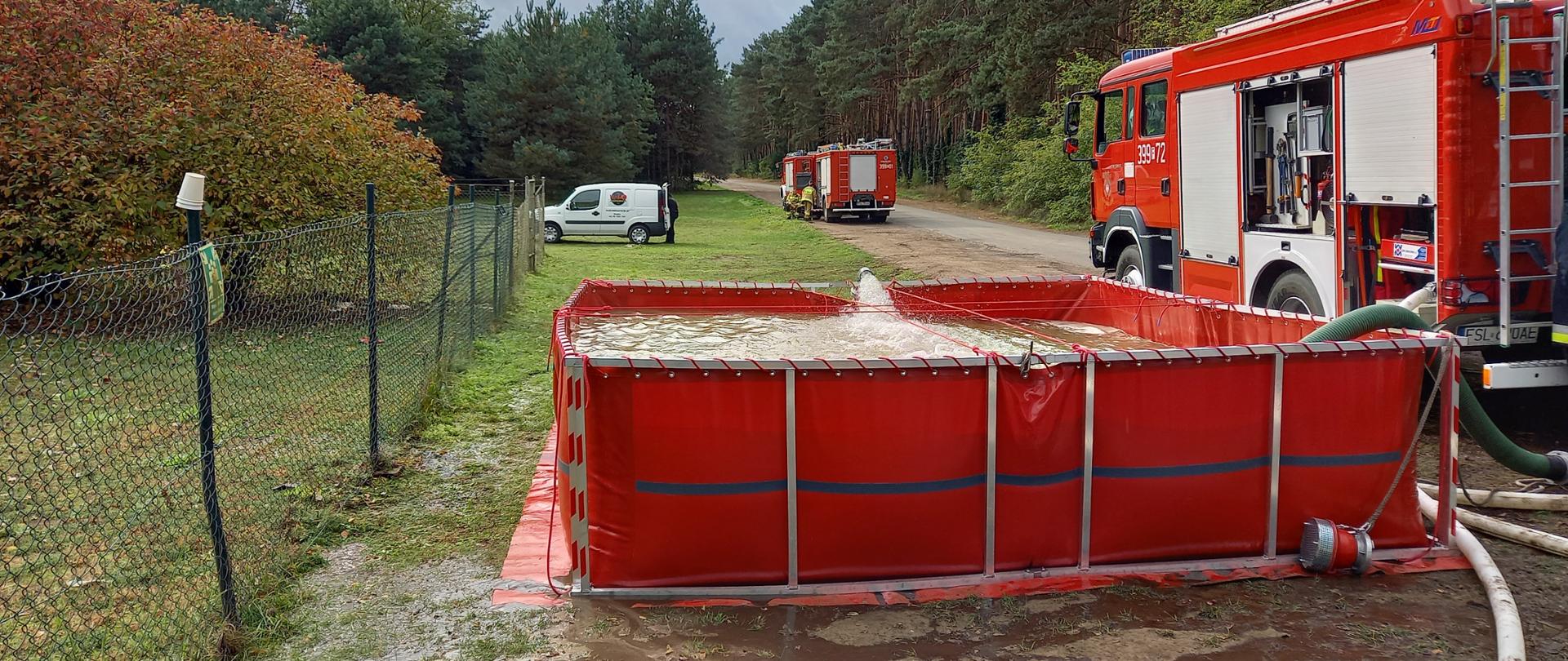 Przewoźny zbiornik strażacki napełniony wodą. Z jego prawej strony samochód pożarniczy z rozwiniętą linią ssawną do zbiornika. W tle kolejne samochody pożarnicze.