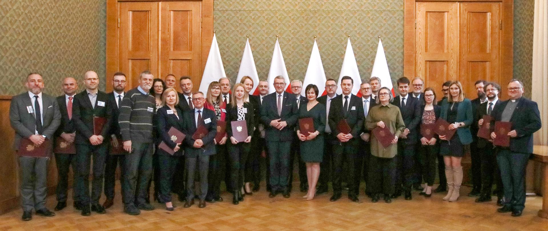 Zdjęcie zbiorowe, na drewnianej podłodze tle zielonej ściany wyłożonej drewnianymi panelami stoi duża grupa elegancko ubranych osób, trzymają w rękach czerwone teczki ze złotym godłem.