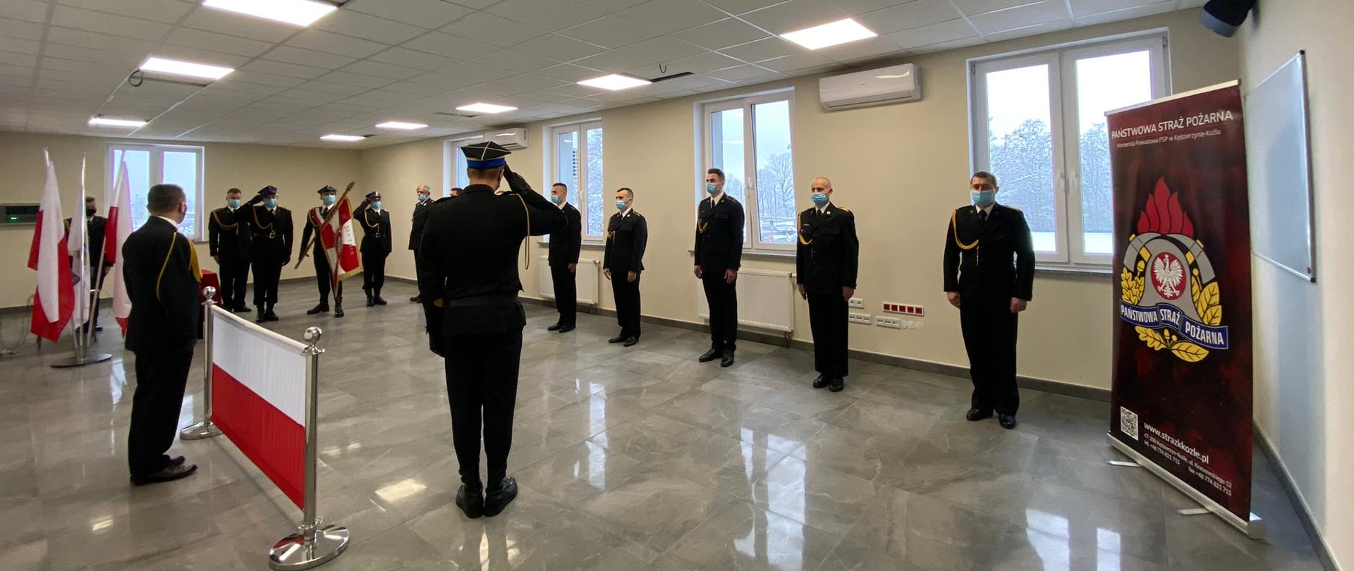 Strażacy stoją w rzędzie po lewej stronie flaga Polski oraz poczet sztandarowy ze sztandarem
