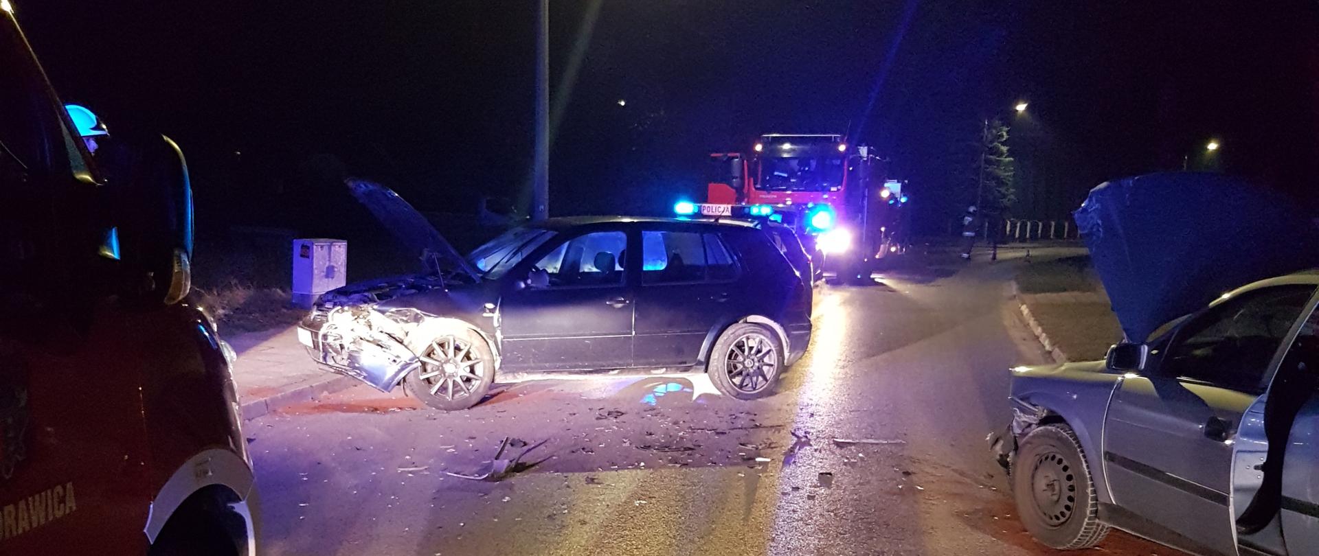 Zdjęcie przedstawia miejsce wypadku - na środku drogi znajdują się rozbite dwa pojazdy: po lewej VW Golf, po prawej Opel Astra. W tle widać samochód pożarniczy. 