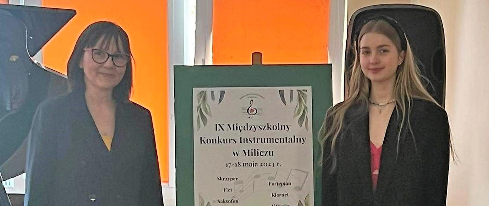 dwie osoby stojące na scenie obok fortepianu, między nimi sztaluga z plakatem konkursu instrumentalnego