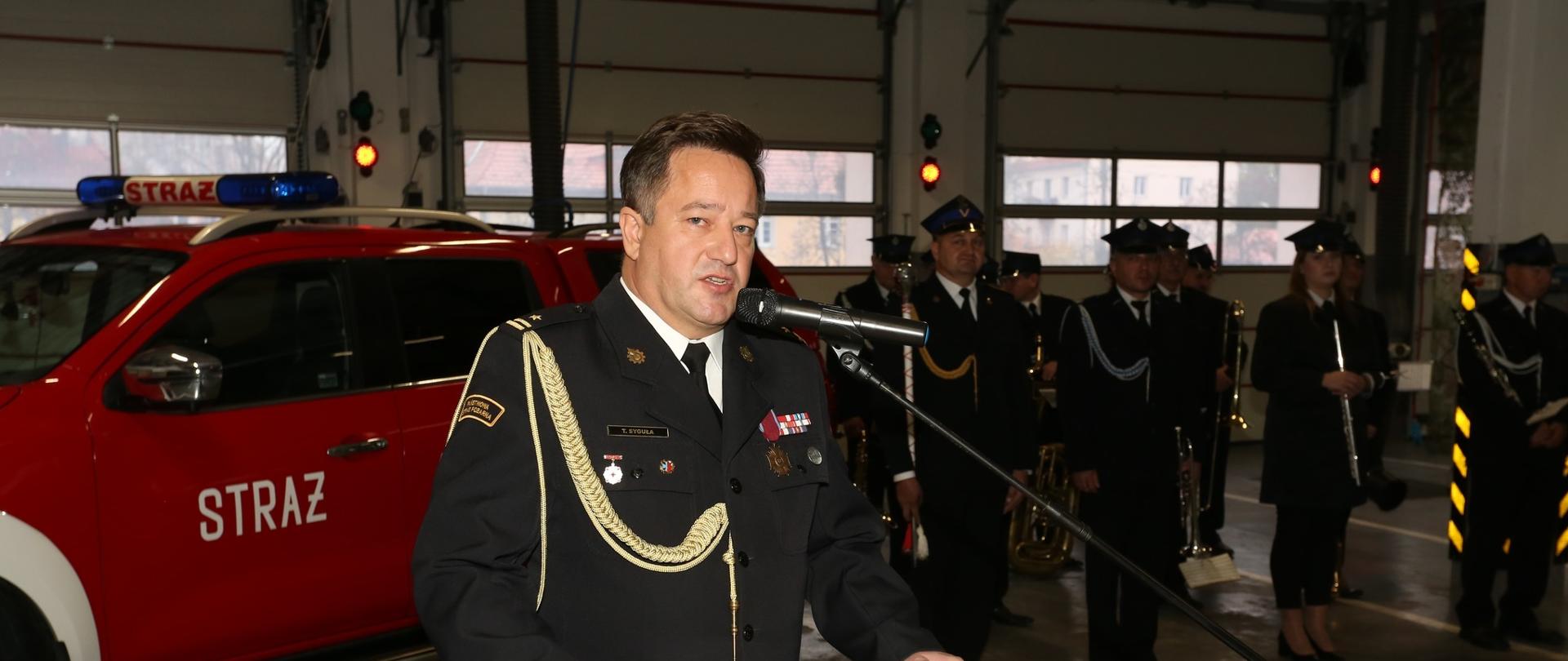 Zdjęcie zrobione na garażu JRG w Kętrzynie w tle czerwony wóz z niebieskimi kogutami. Funkcjonariusze ubrani w czarny mundur galowy, z żółtym sznurem przypiętym od pagonu z lewej strony do pierwszego guzika. Przy mównicy stoi nowopowołany Komendant Powiatowy.