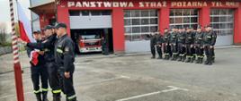 Kolorowe zdjęcie wykonane na zewnątrz, w jasny dzień, przedstawia uroczystą zbiórkę strażaków w dwuszeregu na placu przed budynkiem Państwowej Straży Pożarnej. Trzech strażaków podnosi Flagę Polski na maszt. Pozostali strażacy zwróceni frontem do masztu flagowego stoją na baczność a dowódca salutuje. W tle czerwona ściana frontowa budynku strażnicy z bramami garażowymi. Jedna z bram podniesiona, w której widać samochód pożarniczy i stojącego strażaka. Na ścianie widoczny srebrny napis Państwowa Straż Pożarna tel. 998. 