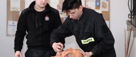 na stole leży fantom do nauki intubacji ratownik osp ubrany w ubranie koszarowe zakłada manekinowi rurkę gardłowo-ustną drugi ratownik ubrany w ubranie koszarowe przygląda się koledze