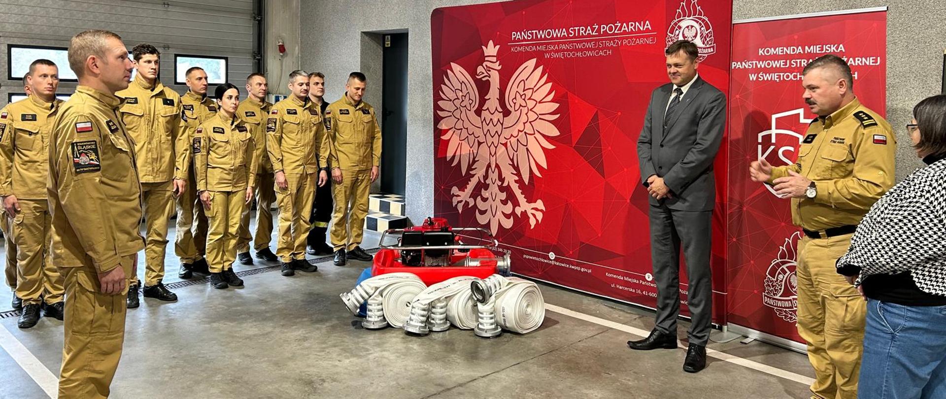 Na zdjęciu widzimy uroczyste wydarzenie z udziałem funkcjonariuszy Państwowej Straży Pożarnej. Grupa strażaków w piaskowych mundurach stoi w szyku w garażu. Na rękawach mają widoczne emblematy, a ich postawa sugeruje, że uczestniczą w ceremonii wręczenia sprzętu.