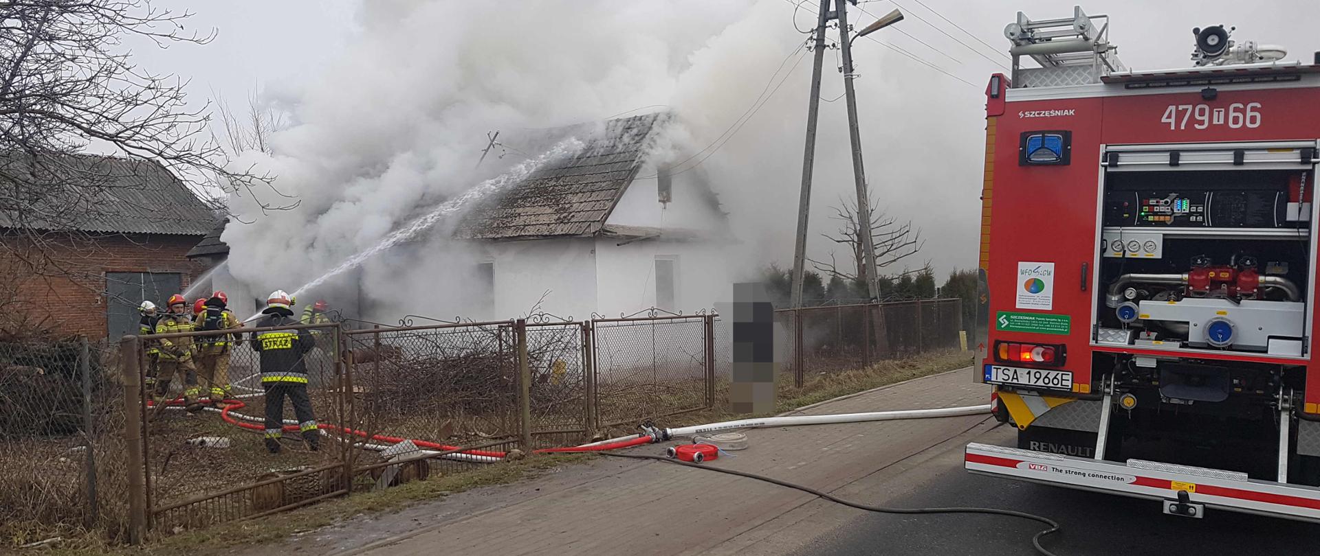 Zdjęcie zrobione w dzień. Na zdjęciu widać palący się budynek mieszkalny i dużo dymu. Zdjęcie przedstawia strażaków, którzy rozwijają węże i prowadzą akcję gaśniczą budynku. W dalszej części zdjęcia widać tył samochodu strażackiego. 