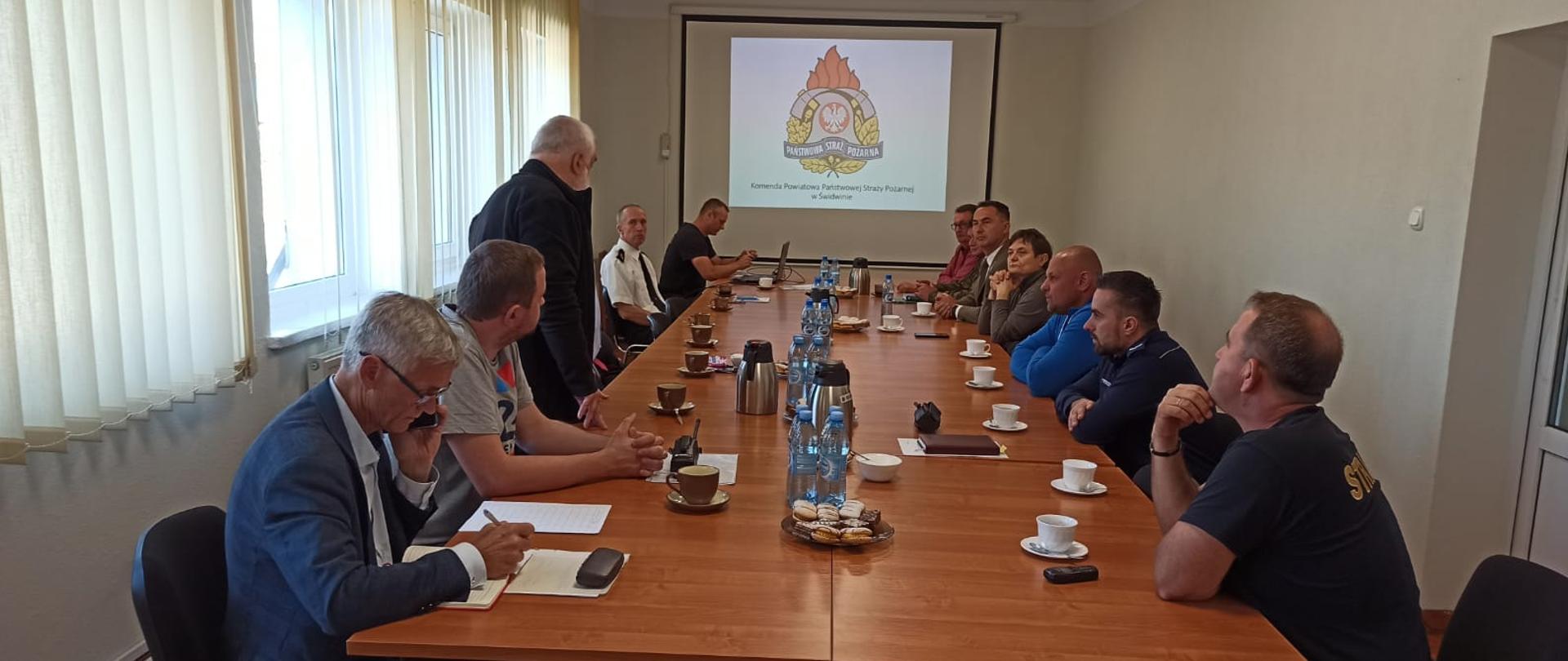 Zdjęcie przedstawia spotkanie, które odbywa się w sali szkoleniowej. W centrum zdjęcia widać stoły przy których siedzą funkcjonariusze oraz inni uczestniczący w szkoleniu. Jena osoba prowadząca szkolenie, która siedzi jest ubrana w niebieską koszulę. Na tle rzutnika widać logo Państwowej Straży Pożarnej.