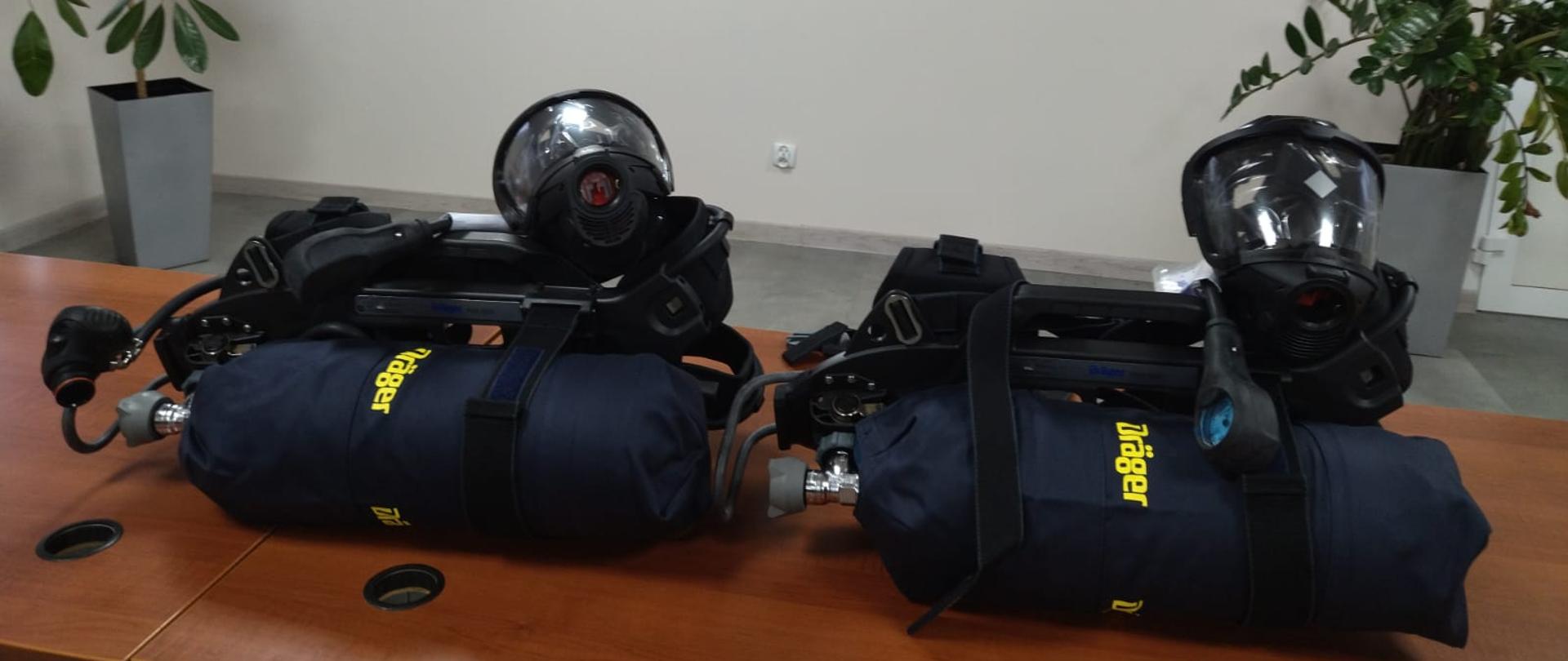 2 komplety aparatów powietrznych lezą na biurkach na świetlicy. W tle ściana z napisem Komenda Powiatowa Państwowej Straży Pożarnej w Makowie Mazowieckim oraz godło Polski i logo PSP.