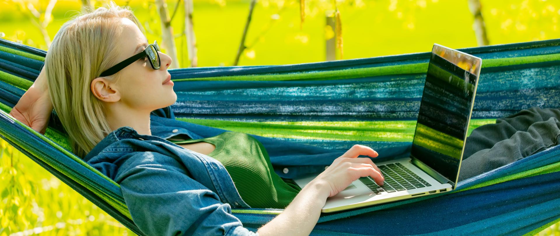 Młoda dziewczyna w okularach przeciwsłonecznych leży na hamaku i korzysta z laptopa.