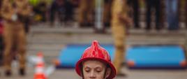 Młoda dziewczyna w czerwonym kasku i koszulce, z determinacją przenosi improwizowanego „manekina” wykonanego ze zwiniętych węży strażackich spiętych taśmą, podczas zawodów lub szkolenia strażackiego. Manekin, przypominający sylwetkę ludzką z ramionami i nogami z węży oraz tułowiem owiniętym srebrną taśmą, jest ciężki i sięga dziewczynie od kolan prawie do ramion, a ona trzyma go oburącz w neonowożółtych rękawicach, opierając go o siebie. Scena odbywa się na brukowanym placu z widocznymi w tle białymi liniami, pachołkami ostrzegawczymi, niebieskimi matami do ćwiczeń oraz zamazanymi sylwetkami innych uczestników i sędziów w mundurach.