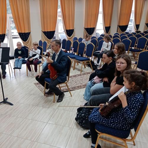 Zdjęcie przedstawia dużą salę z krzesłami ustawionymi w rzędach, oraz częściowo skupionymi wokół mężczyzny ze skrzypcami na których siedzą dzieci obserwujące wydarzenie. Naprzeciw mężczyzny, ubranego w garnitur, noszącego okulary, tyłem stoi dziewczynka o blond włosach splecionych w warkocz, ubrana w dres w ciemnym kolorze, przed nią stoi czarny pulpit z nutami.