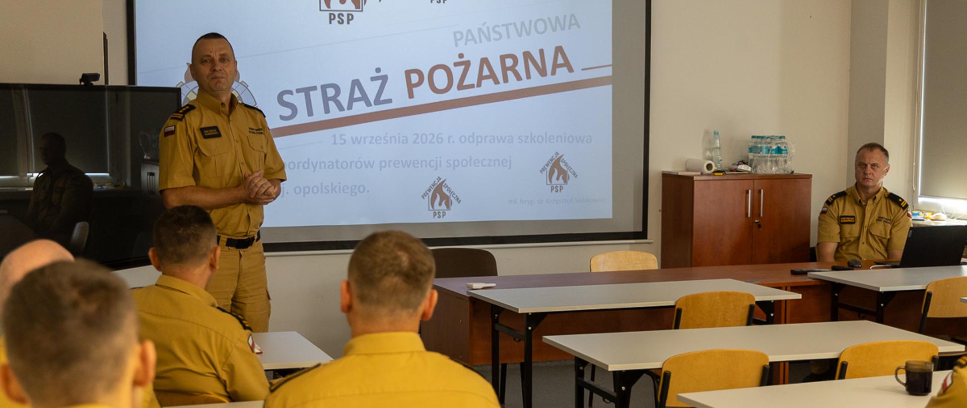 Kilku strażaków PSP w żółtych mundurach siedzi w rzędach przy biurkach, zwróconych w stronę instruktora stojącego z przodu sali. Na plecach mundurów widoczny jest napis „Państwowa Straż Pożarna”. Instruktor prezentuje treści wyświetlane na dużym ekranie z interfejsem komputerowym. Sala jest dobrze oświetlona, z białymi ścianami i sufitem z paneli.