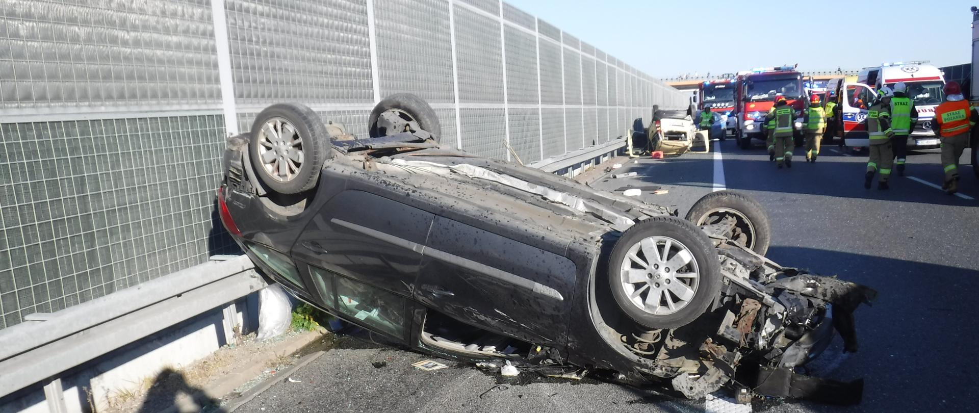 Zdjęcie przedstawia przewrócony na dach samochód osobowe stojące na pasie awaryjnym autostardy A4 w kierunku Krakowa, tył pojazdu opary jest na barierce za pojazdem znajdują się pororzucane cześci pojazdu z z tył pokazdu znajduje się barireki dziękochłonne. Na pojazdem pare metrów widać drugi pojazd w kolorze zółtym wyrócony na dach. obok złótego pojazdu po prawej stronie znajdują się dwa czerwone pojazdy strazy pożarnej z włączonymi światłami błyskowymi barwy niebieskiej i jeden pojazd Zespołu Ratownictwa Medycznego. Obok pojazdów znajduja się pieciu strażaków ubranych w piaskowe mundury i czerwone hełmy.Jeden z nich dodatkowo ma pomarańczową kamizelkę Warunki atmosferyczne bardzo dobre słoneczna pogoda.