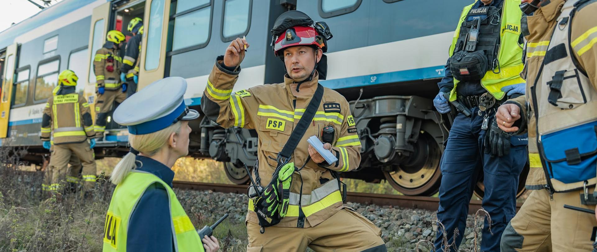 Na zdjęciu znajdują się strażacy oraz policjanci w tle wagon pociągu pasażerskiego 