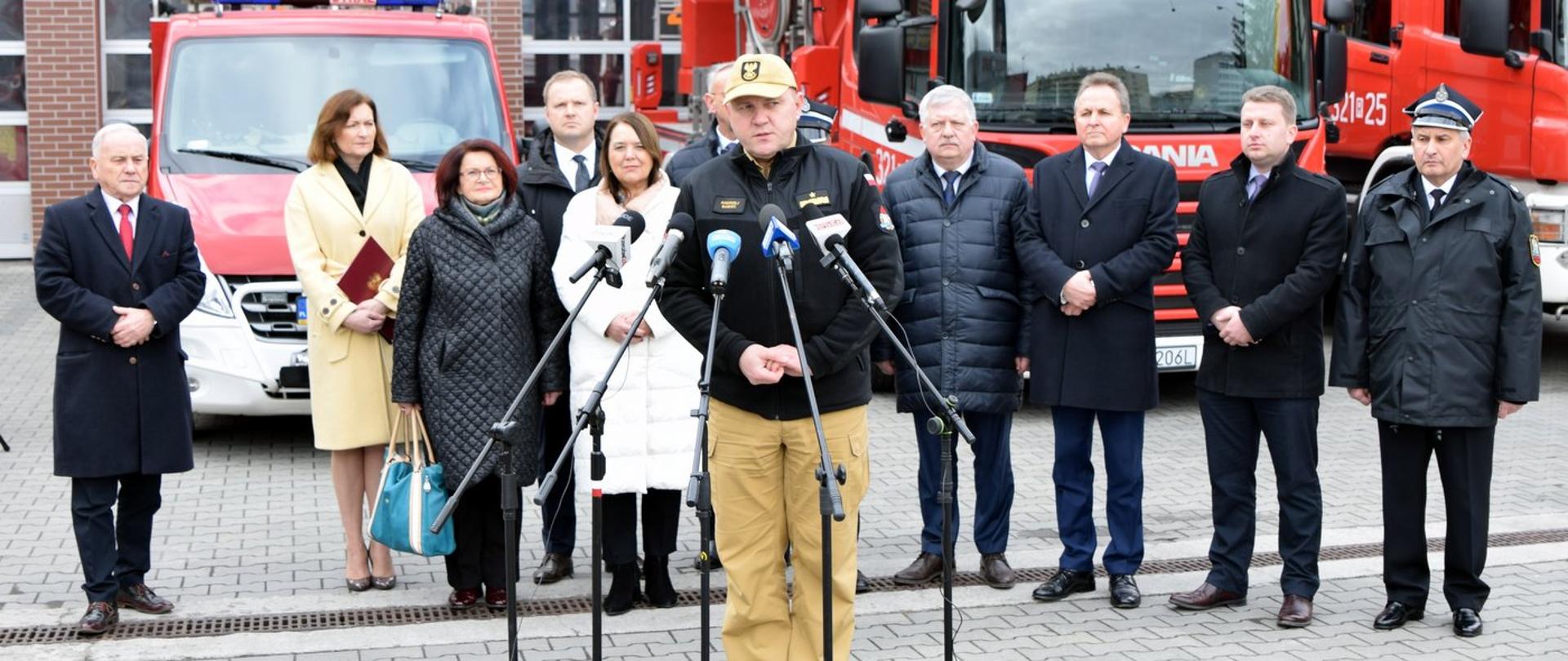 Zdjęcie zrobione na zewnątrz, przed garażami Państwowej Straży Pożarnej. Fotografia przedstawia uczestników briefingu prasowego. Przed mikrofonami stoi oficer PSP w stopniu generała i przemawia. Za nimustawieni w szeregu stoją pozostali uczestnicy briefingu prasowego. Za zgromadzonymi osobami stoją trzy samochody ratowniczo-gaśnicze.