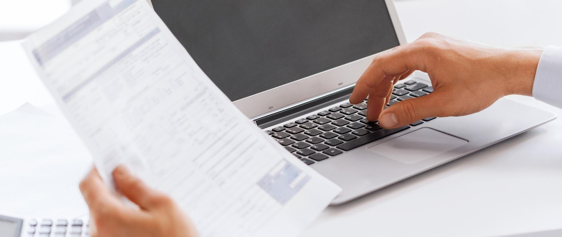 Dłoń trzymająca papier, druga dłoń na klawiaturze komputera.