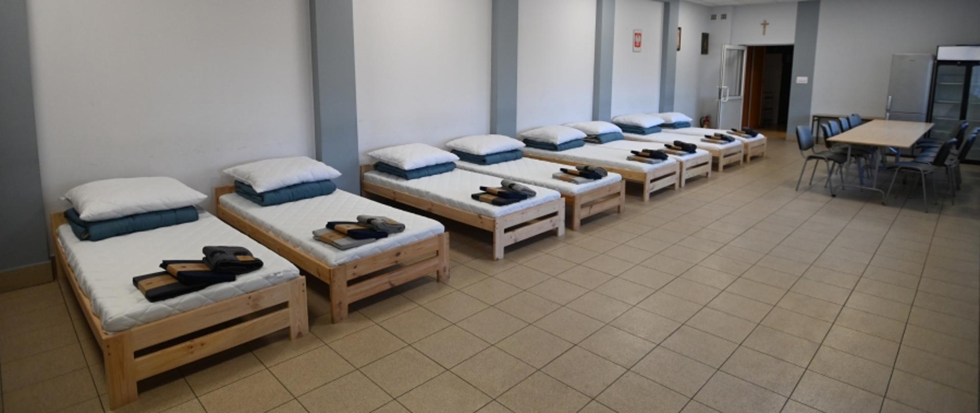 Na zdjęciu widoczne siedem łózek na sali. Łóżka drewniane, z białymi materacami, na łóżkach komplet pościeli.