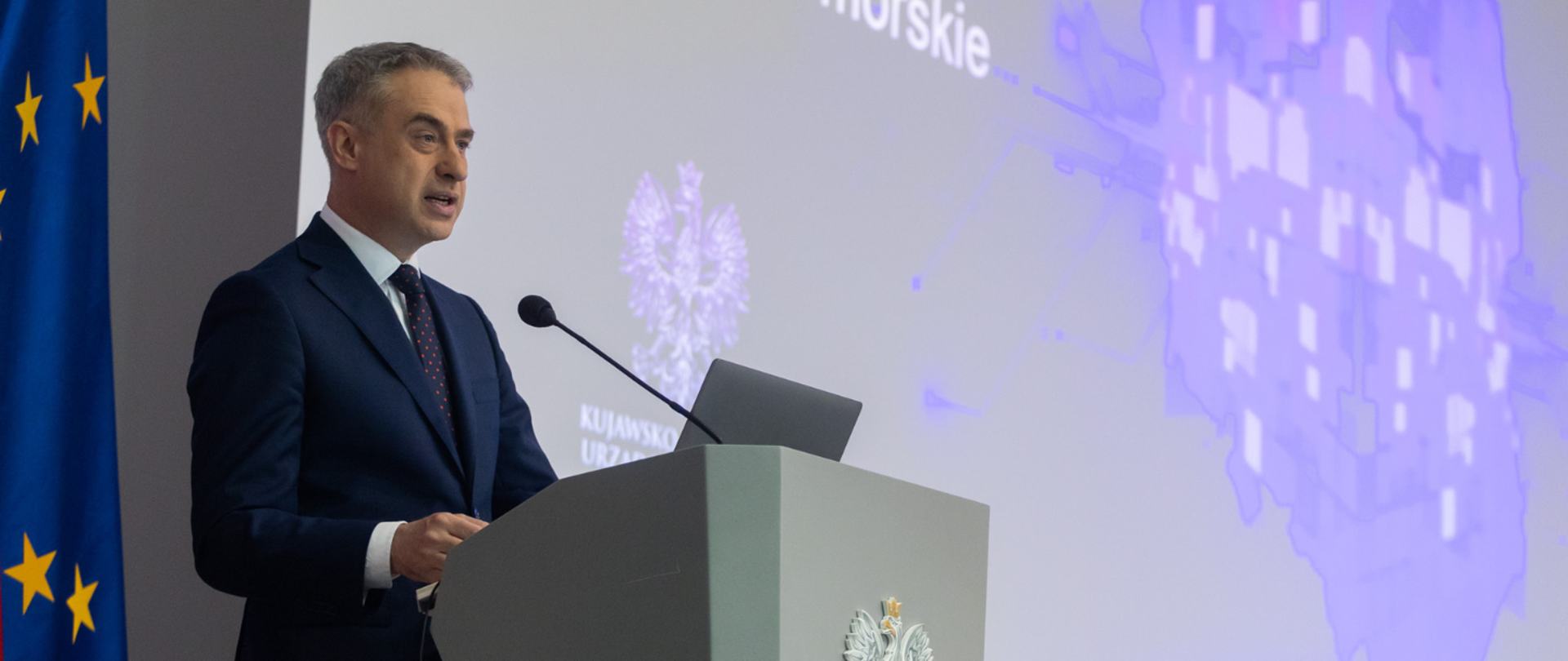 minister cyfryzacji Krzysztof Gawkowski stoi przy mównicy, za nim widać slajd prezentacji. 