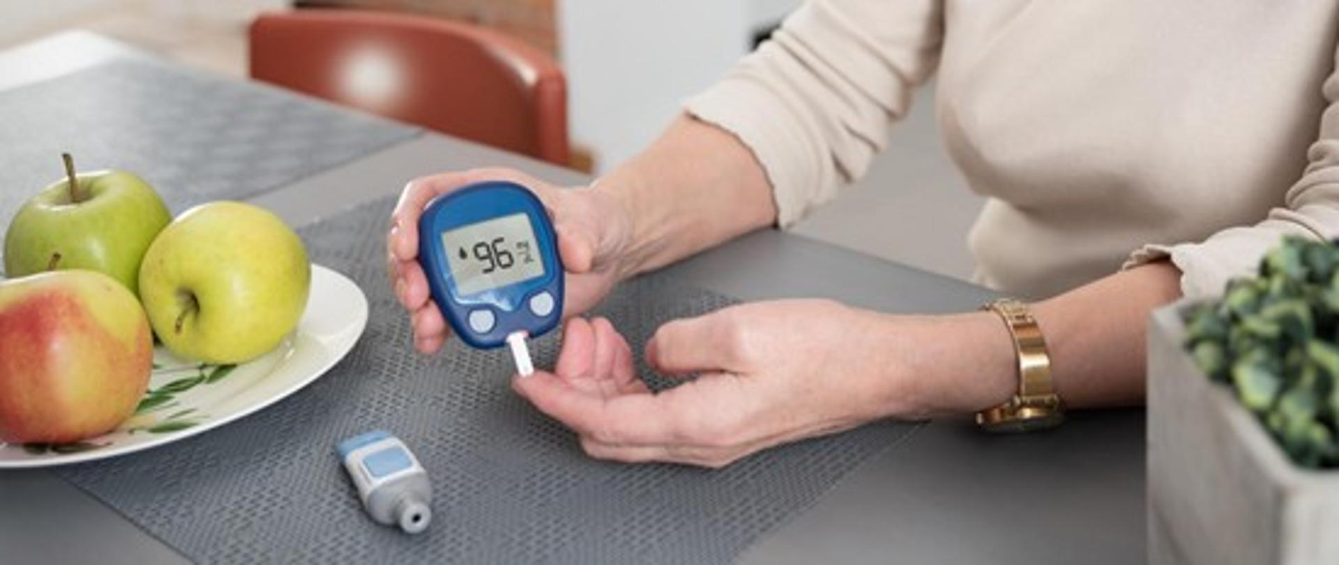 Osoba siedząca przy stole i mierząca gleukometrem poziom cukru z palca wskazującego. 