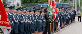 Szereg funkcjonariuszy Krajowej Administracji Skarbowej w galowych mundurach stoi w poczcie sztandarowym obok flag państwowych i związkowych. Obok nich zgromadzeni goście i przedstawiciele innych formacji.
