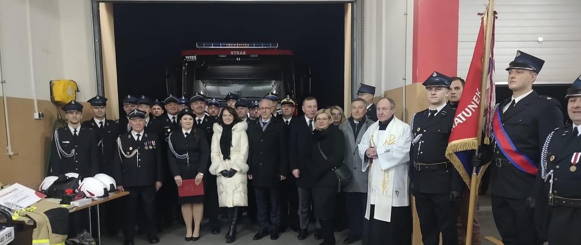 Na tle samochodu strażackiego zbiórka zebranych gości wraz ze strażakami w mundurach