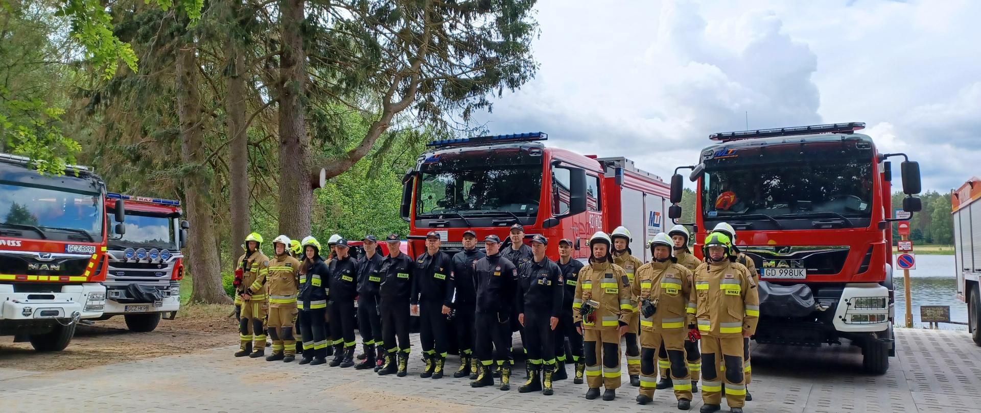 Zdjęci grupowe. Na pierwszym planie widać strażaków stojących w szeregu na tle samochodów pożarniczych. W tle jezioro i punkt czerpania wody.