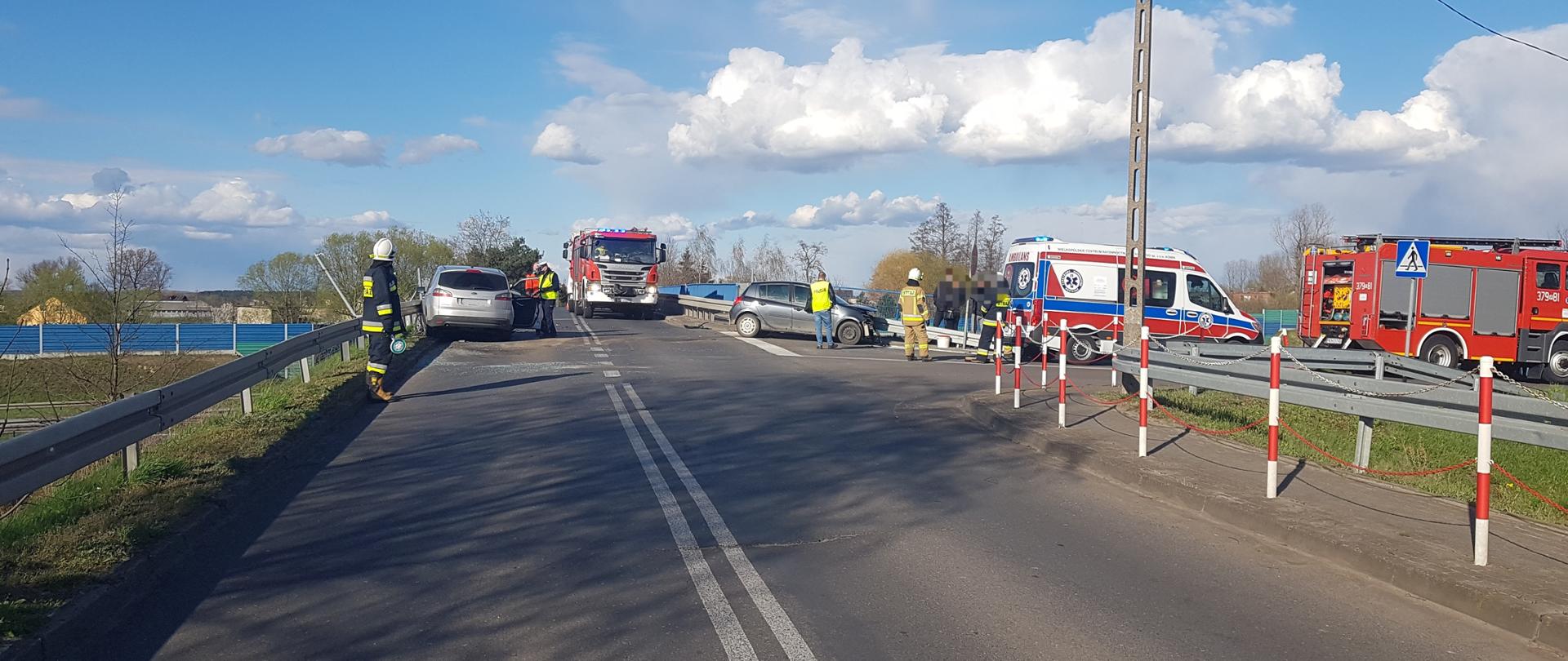 Zdjęcie przedstawia dwa samochody osobowe , które się zderzyły. Samochód koloru srebrnego stoi tyłem, natomiast samochód koloru ciemnego bokiem. Na zdjęciu widać również samochody strażackie , karetkę pogotowia ratunkowego oraz strażaków i policjantów. 