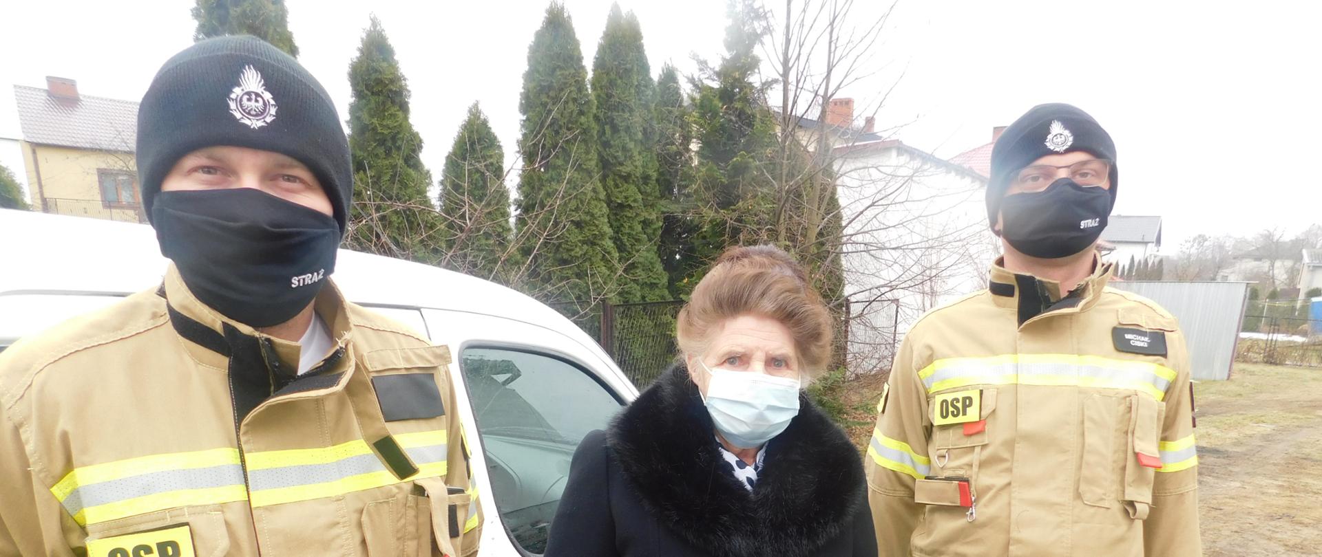 Dwóch strażaków i kobieta stoją przed białym samochodem. Kobieta stoi pomiędzy strażakami.