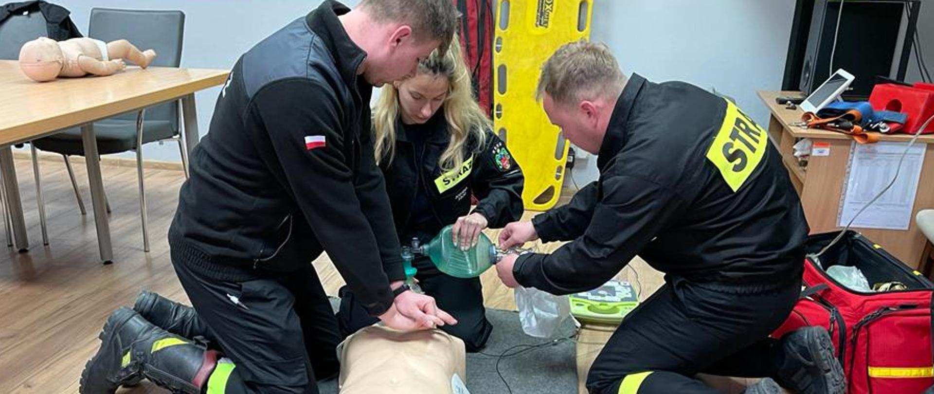 trzech strażaków w czasie egzaminu. Prowadzą czynności z zakresu kwalifikowanej pierwszej pomocy na manekinie