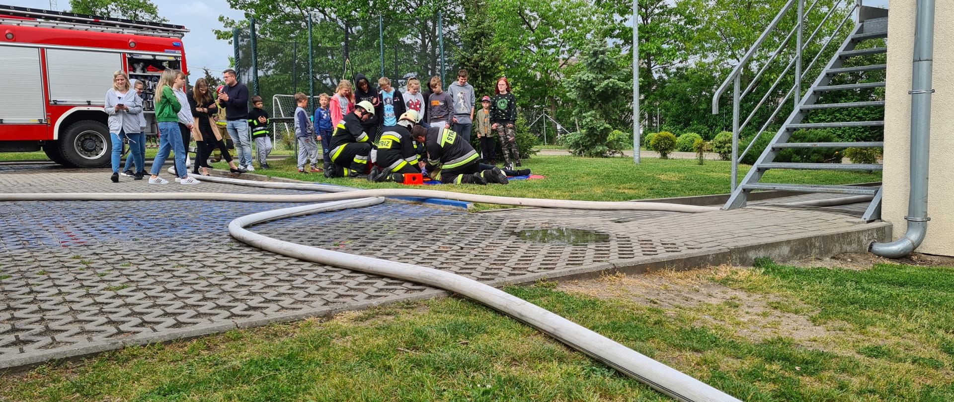 zdjęcie wykonane w dzień. Przedstawia grupę dzieci podczas ćwiczeń strażackich. Dzieci przyglądają się strażakom, którzy udzielają pomocy osobie poszkodowanej. W tle samochód strażacki. Po prawej stronie budynek. Strażacy w umundurowaniu specjalnym bokiem na zdjęciu. 