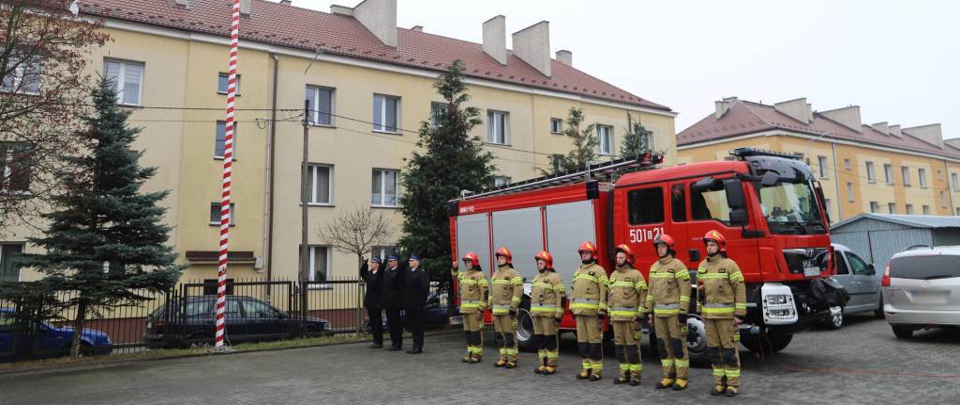 Zdjęcie przedstawia poczet sztandarowy ubrany w mundury wyjściowe w kurtce zimowej z salutującym dowódcą pocztu oraz strażaków z podziału bojowego Jednostki Ratowniczo-Gaśniczej Komendy Powiatowej Państwowej Straży Pożarnej w Zambrowie ubranych w ubranie bojowe w hełmach z salutującym dowódcą zmiany. Strażacy ustawieni w szeregu na placu Komendy Powiatowej Państwowej Straży Pożarnej w Zambrowie, w tle widać samochód bojowy. Zdjęcie wykonano w pochmurny, jesienny dzień. 